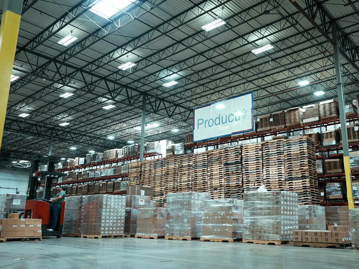 Inside a Productiv warehouse: high-bay racking, staged pallets, a reach truck in motion, and the Productiv sign overhead